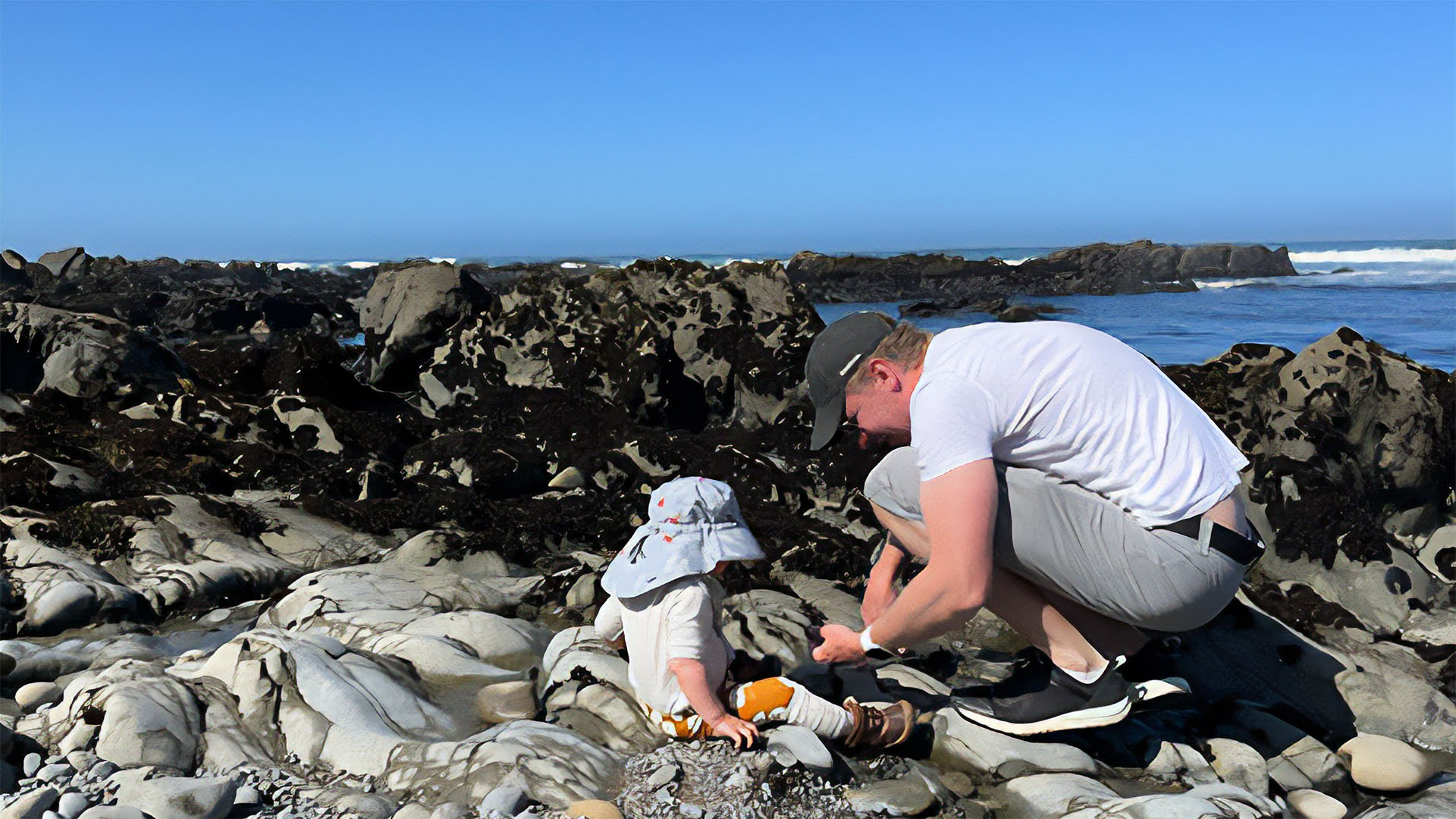 A young toddler and man both wearing hats sit amongst rocks by the ocean with a clear blue sky above them