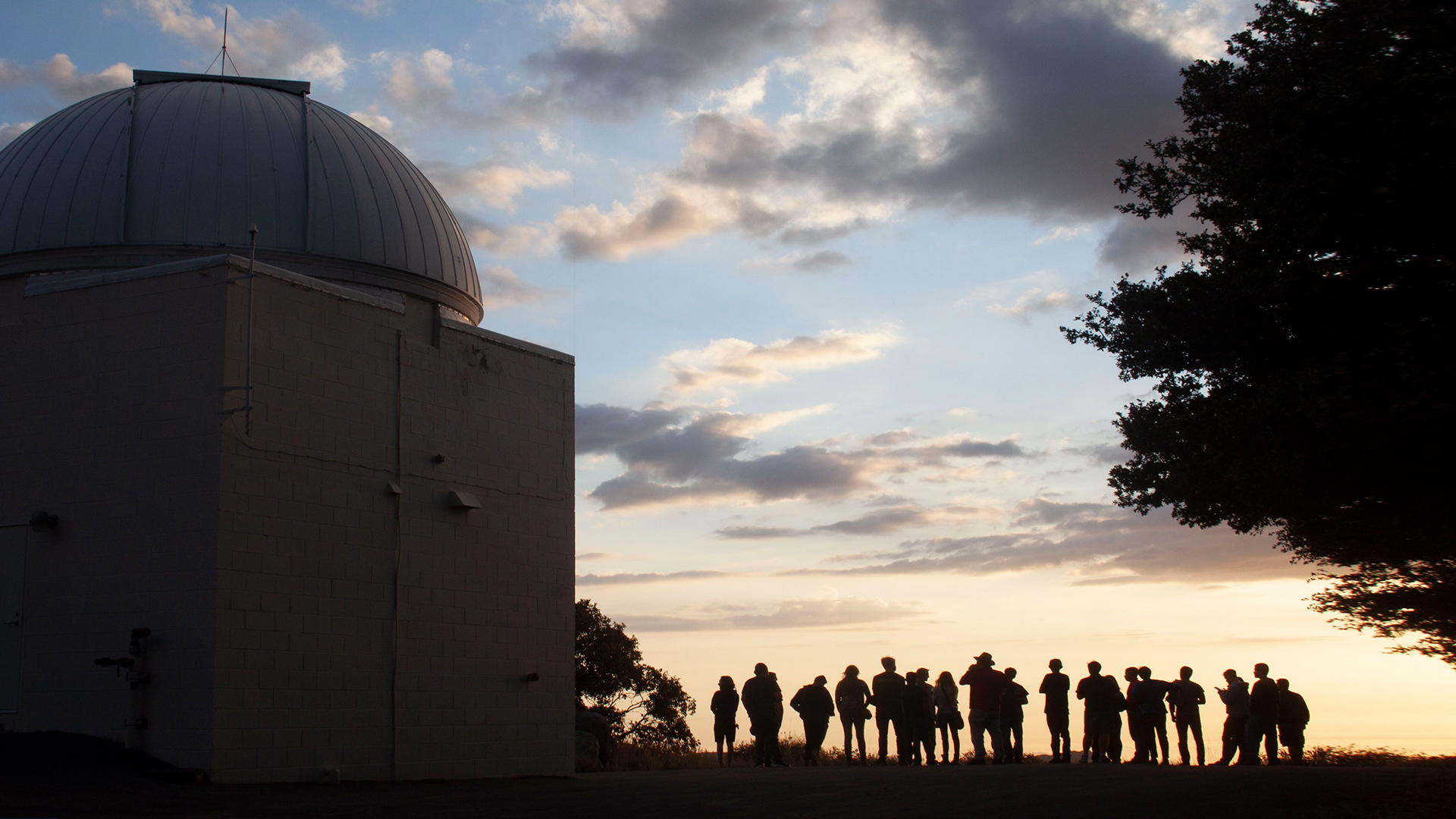At sunset on top of Mt. Laguna Observatory, the silhouettes of 20 people standing next to a telescope dome structure outside.