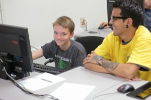kid using computer smiles seated next to student.