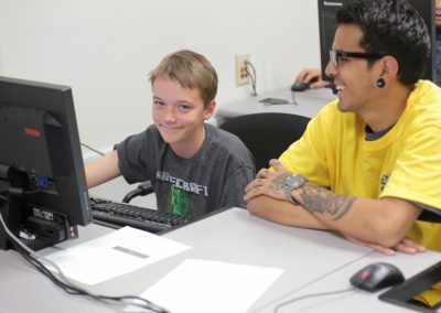 kid using computer smiles seated next to student.