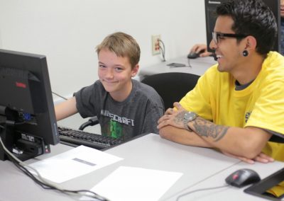 kid using computer smiles seated next to student.