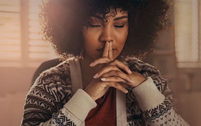 Young woman sitting with hands interlaced with fingers over lips and eyes closed in deep thought.