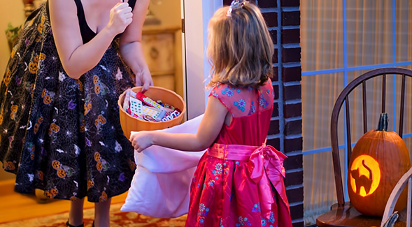 little girl in dress standing at door holding bag with adult handing her candy.