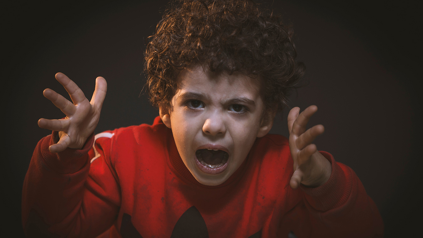 Close-up of young angry boy facing us with curly hair and red sweatshirt gesturing with hands.