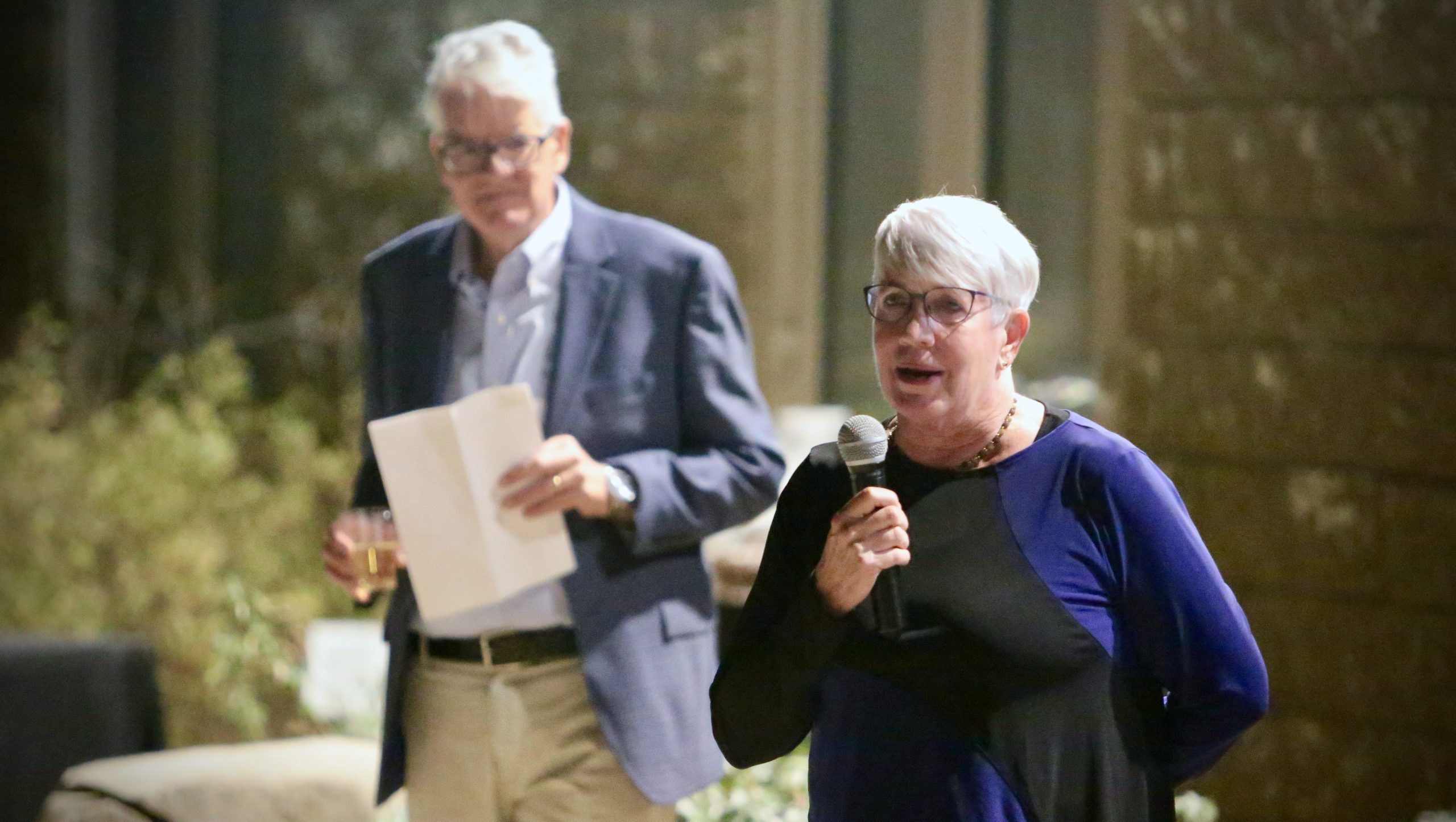 Cathie Atkins, a woman with short grey hair, wearing a blue and black sweater, speaks into a microphone. Dean Roberts, a white-haired man in a blue blazer and khakis stands behind her.