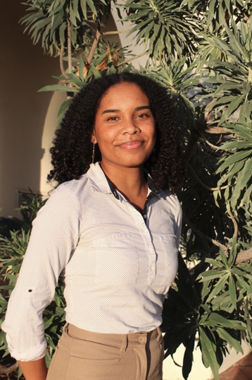 A woman with dark curly hair is smiling during golden hour. She's standing in front of a large tree wearing a white blouse and khaki bottoms.