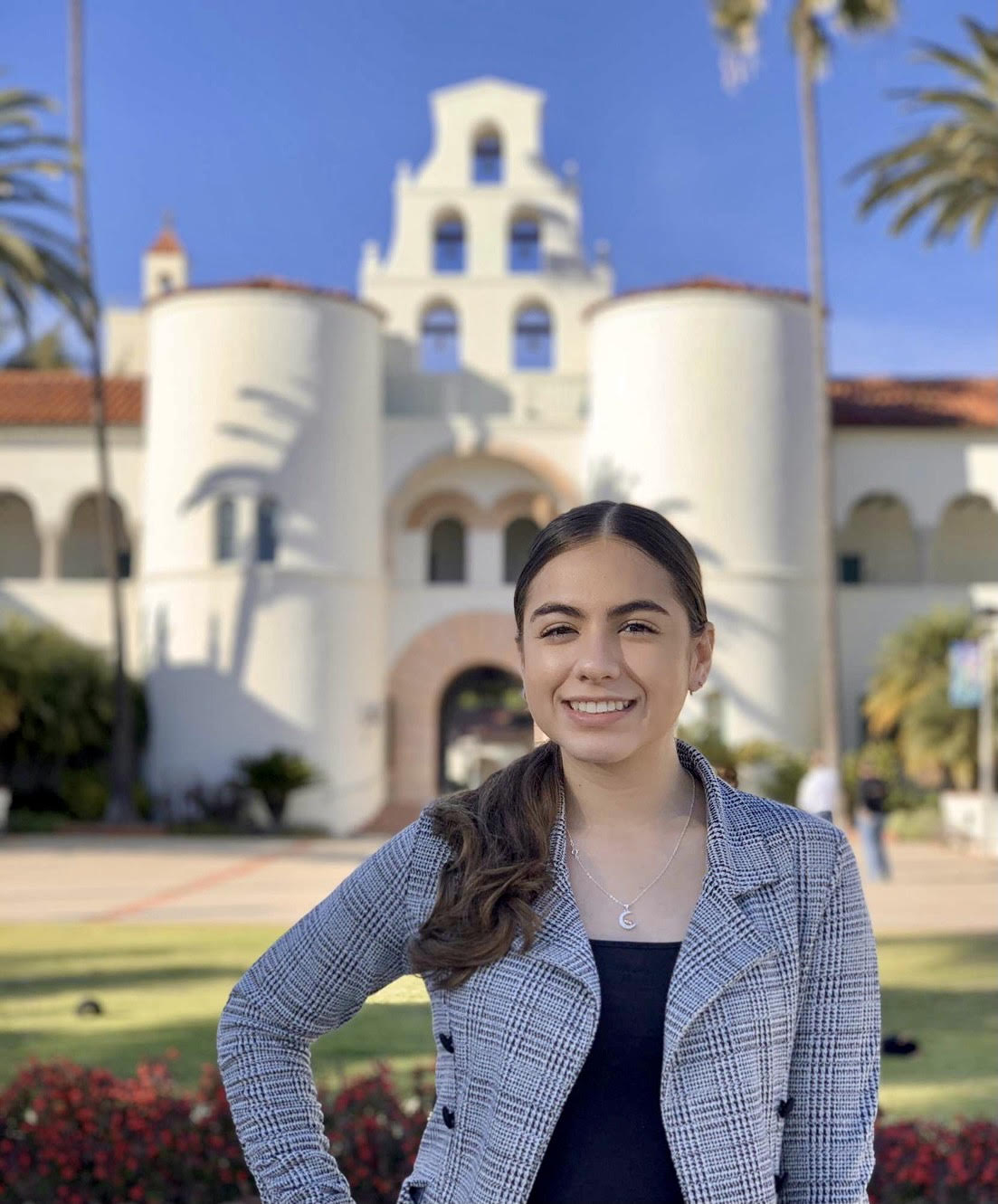 A young woman with dark brown hair stands in front of Hepner Hall. She is wearing a grey plaid blazer and crescent moon necklace