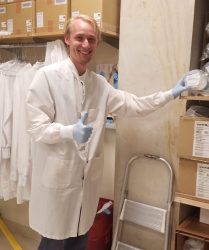 A blonde man in a lab coat smiles in a lab, grabbing lab supplies