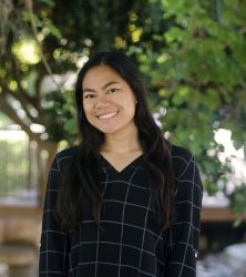 A woman with long dark hair in a long-sleeved black plaid top smiles outside under a tree