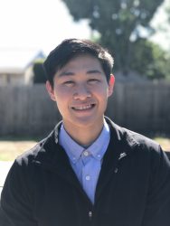 A young Asian man wearing a blue collared shirt and black jacket stands outside, smiling.