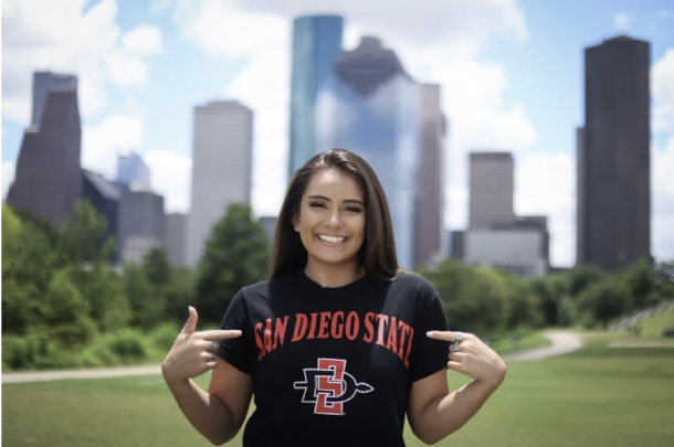 A young woman with dark hair points at her black t-shirt that says San Diego State. A city skyline and park are visible in the background