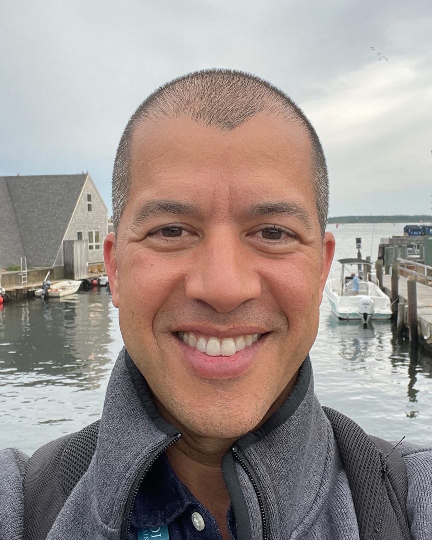 A man smiling wearing a grey sweatshirt with a bay, dock and house visible behind him