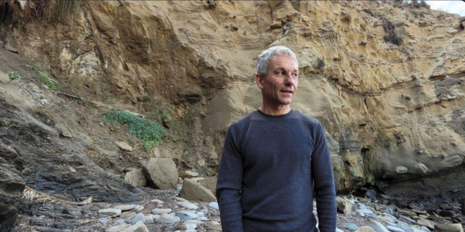 Man in a navy long sleeve shirt stands on a rocky beach looking at the ocean