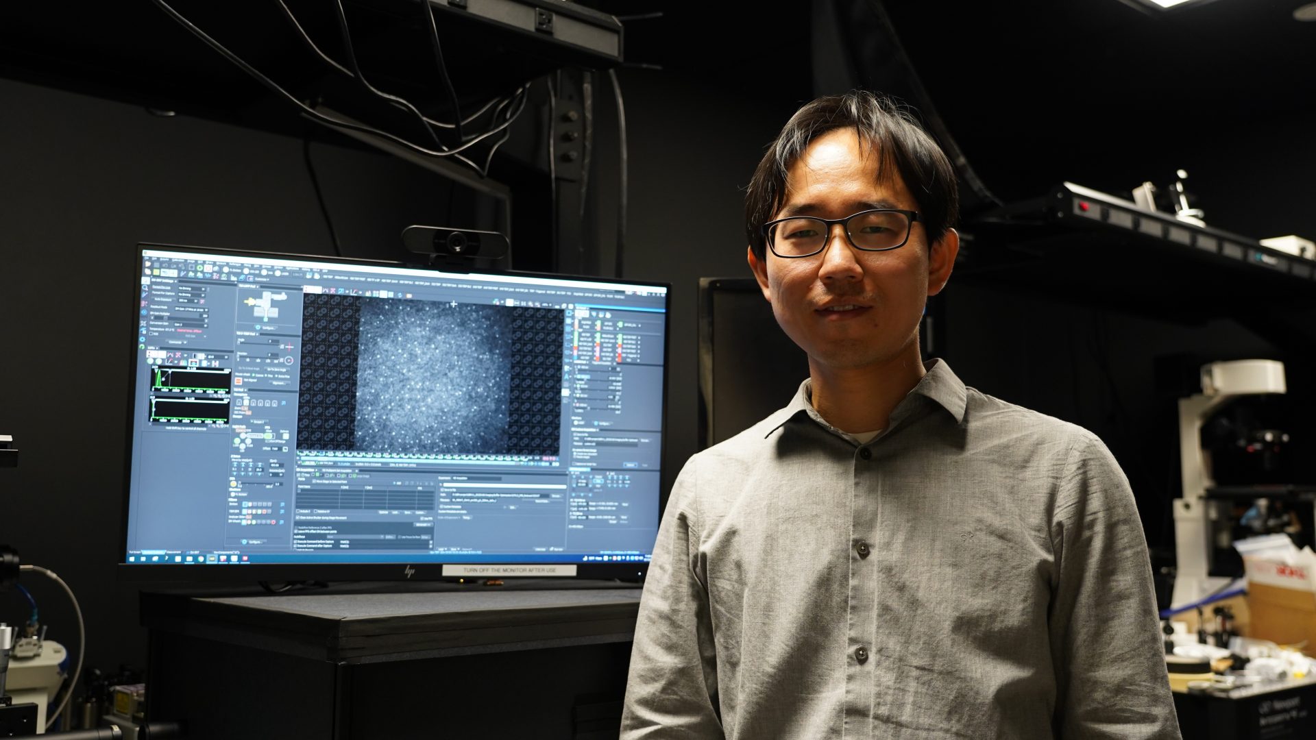Lee stands in his lab, smiling at the camera, with a computer screen visible in the background.