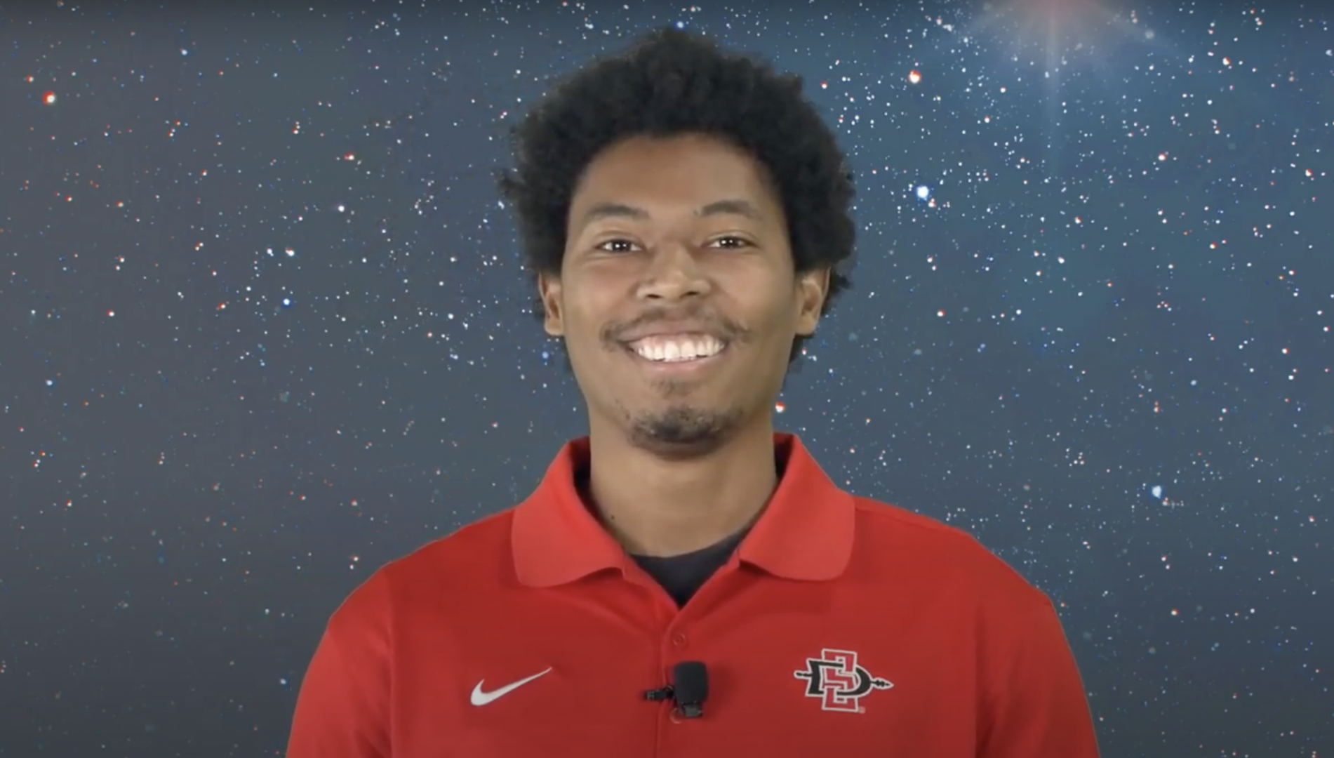 A young man in a red polo shirt smiles in front of a starry virtual background