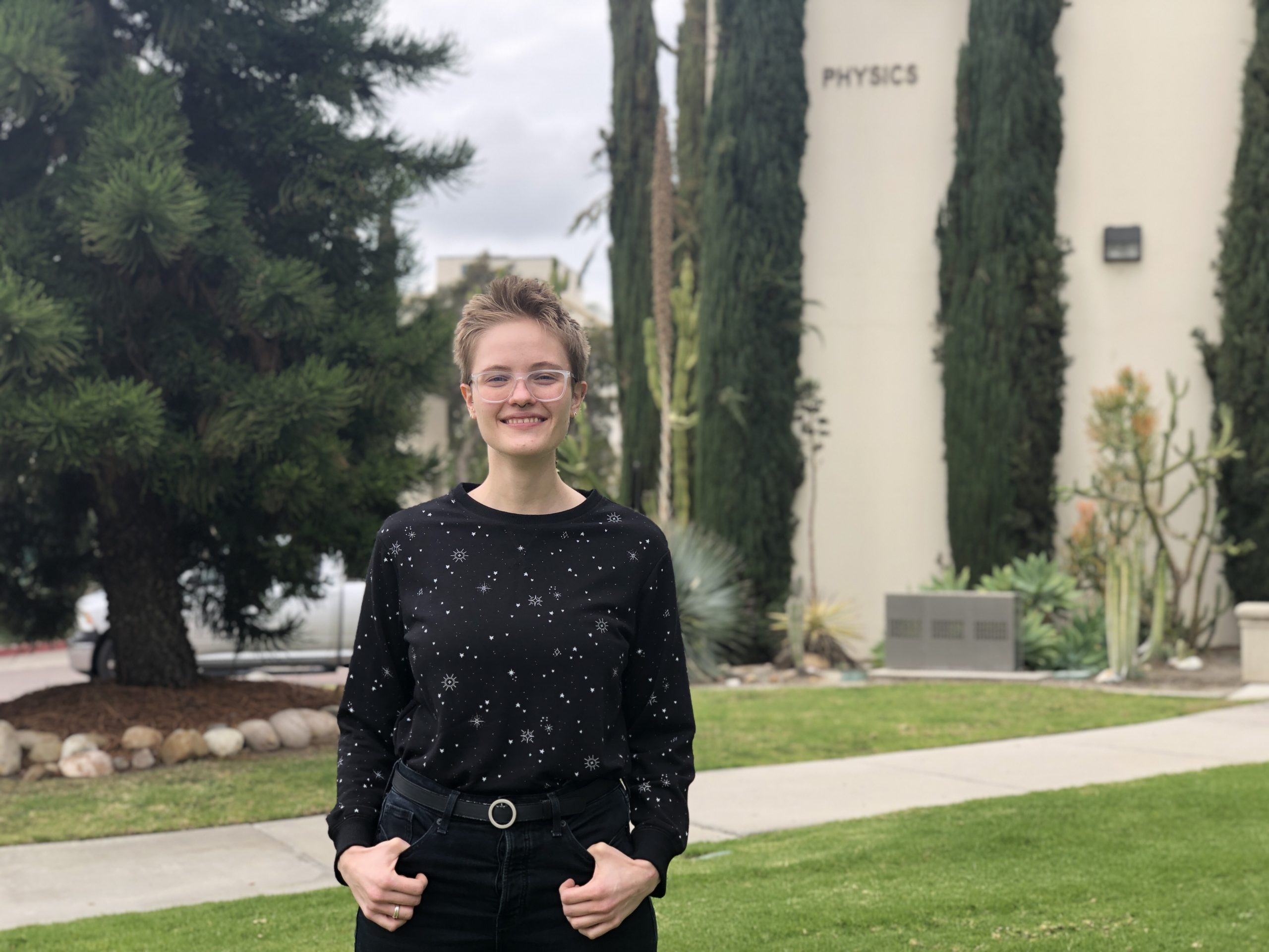 A person wearing a long-sleeve black shirt with a star print and black jeans stands outside of SDSU's physics building