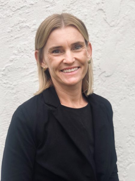 Woman with shoulder-length blonde hair wearing black sweater and shirt smiles in front of white wall