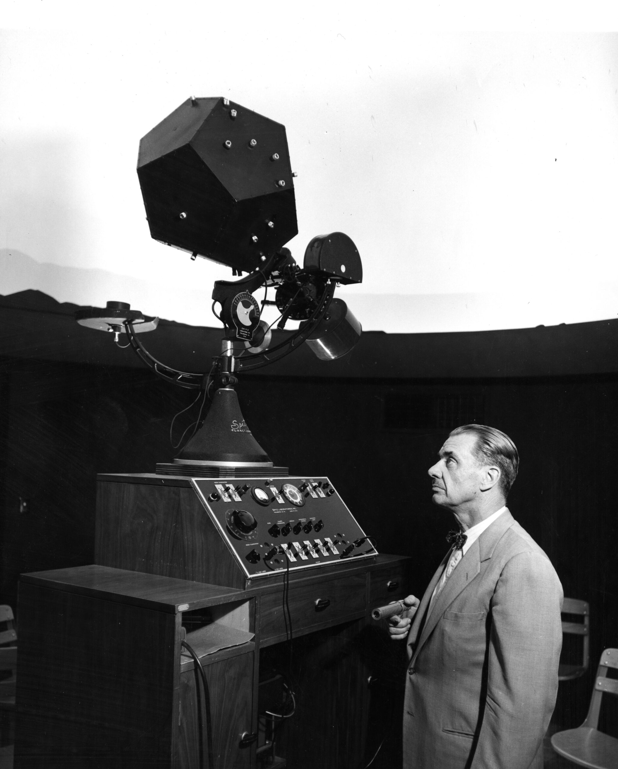 A man in a suit looks at an old-school planetarium projector, greyscale