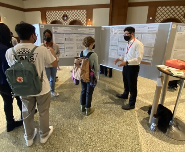 Students wearing masks explain their research to onlookers wearing backpacks and masks.