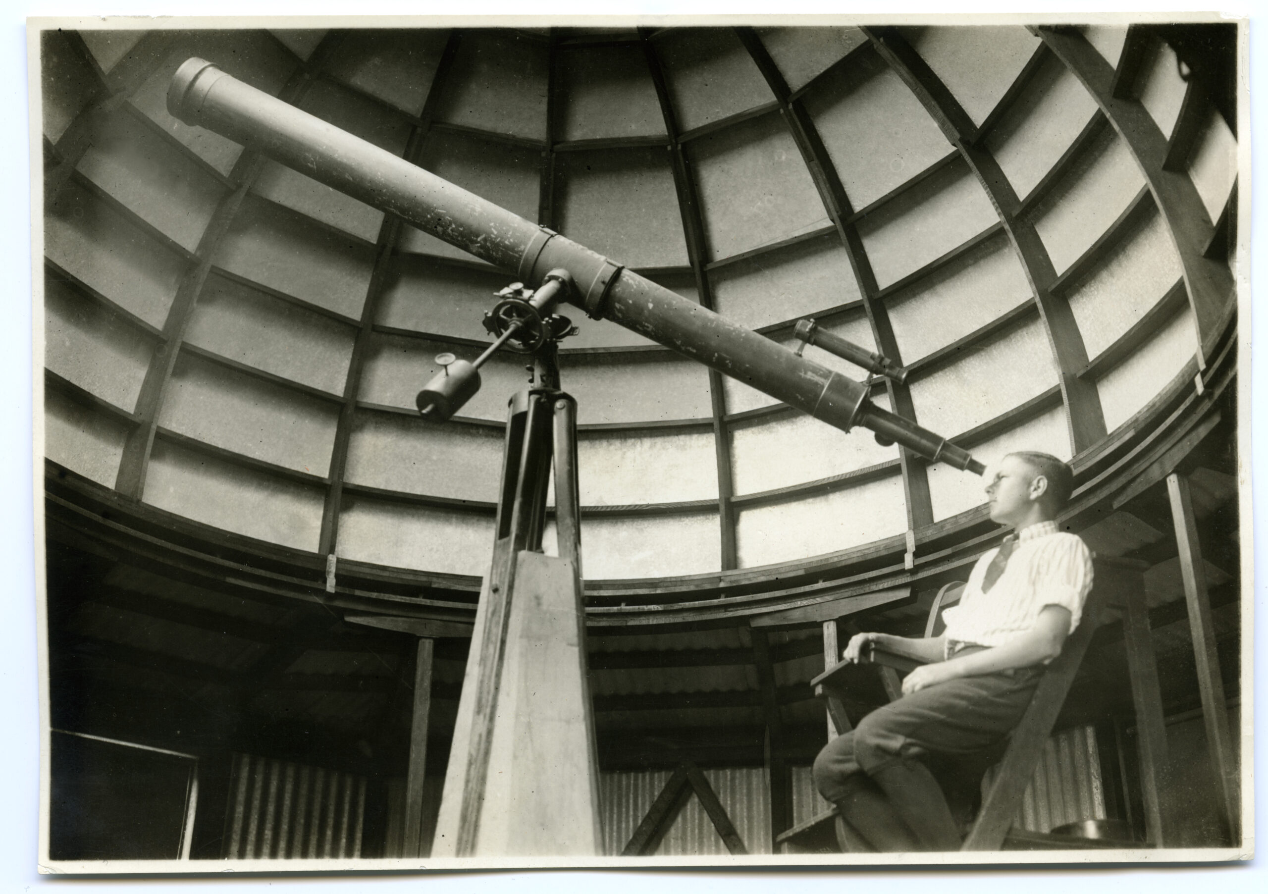 A young man looks through a telescope in a domed building, greyscale