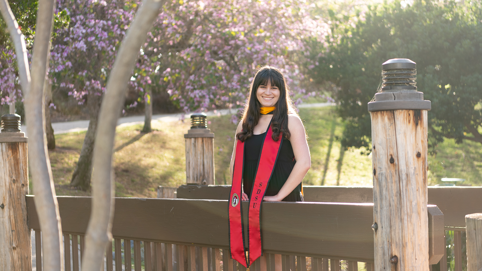 Woman with long dark brown hair wearing a black dress and red graduation sash stands on a bridge during golden hour. A tree with purple blossoms is int he background