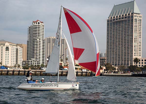 Two people on a boat with a red and white sail on the water in front of a city skyline