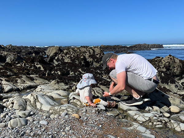 A young toddler and man both wearing hats sit amongst rocks by the ocean with a clear blue sky above them