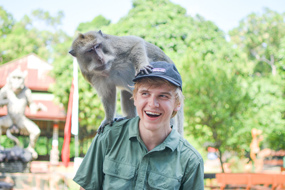 A primate sits on Scott's shoulder while he laughs. Trees and a monkey statue are visible in the background