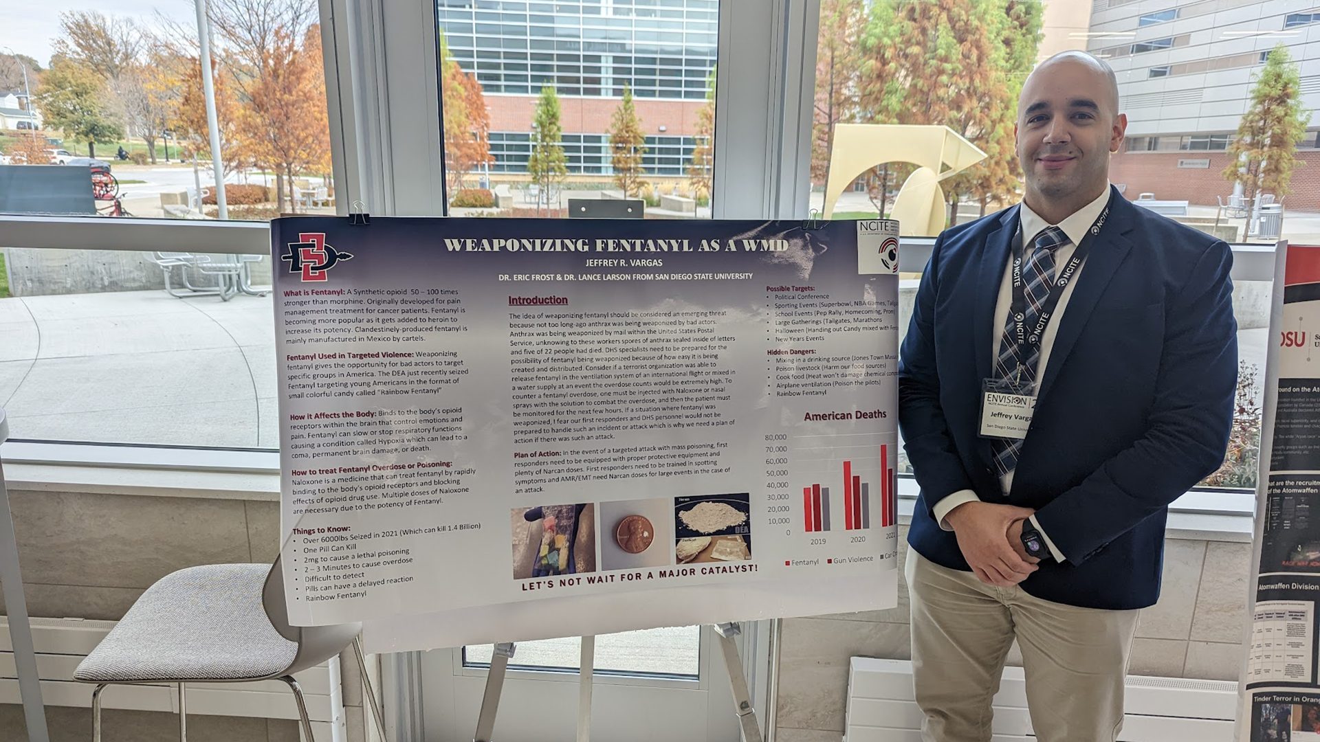 Jeffrey Vargas, dressed in a navy suit, tie and khaki pants, stands next to his poster on fentanyl