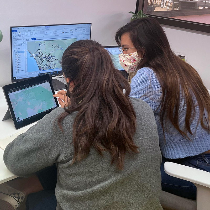 Adriana Rios and colleague sitting at desktop facing computer screens with maps.