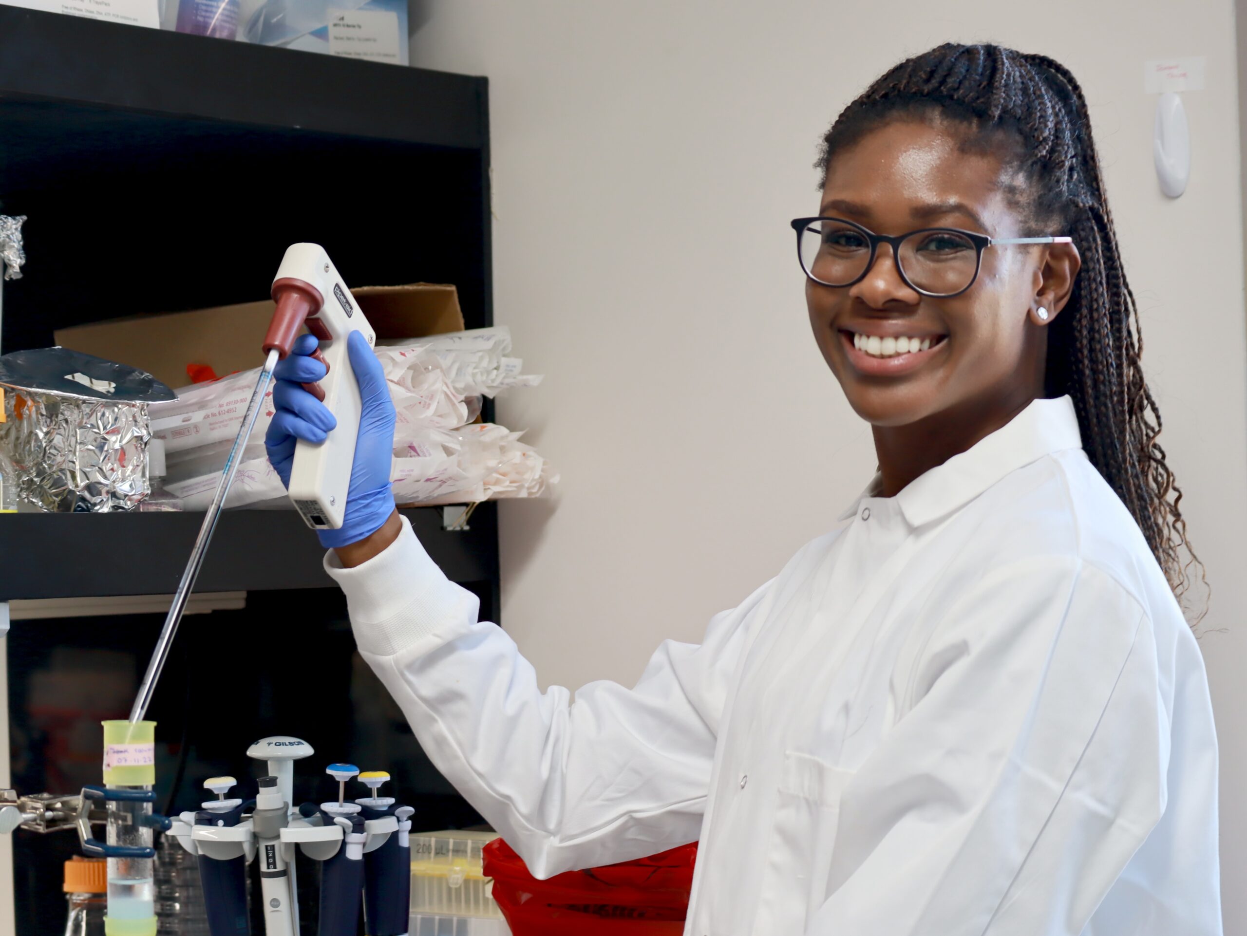 Female student in lab coat smiling while holding a pipette.