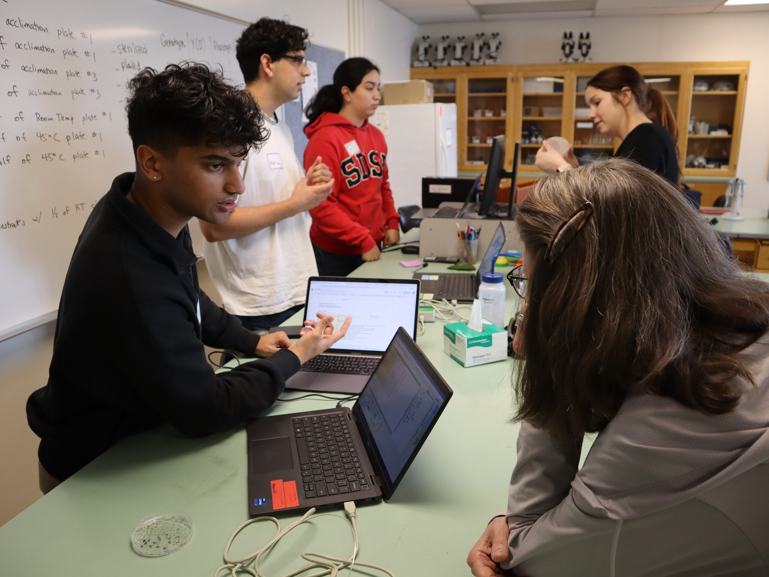 Four students standing around counter with laptops discussing with Dr. Homayouni.