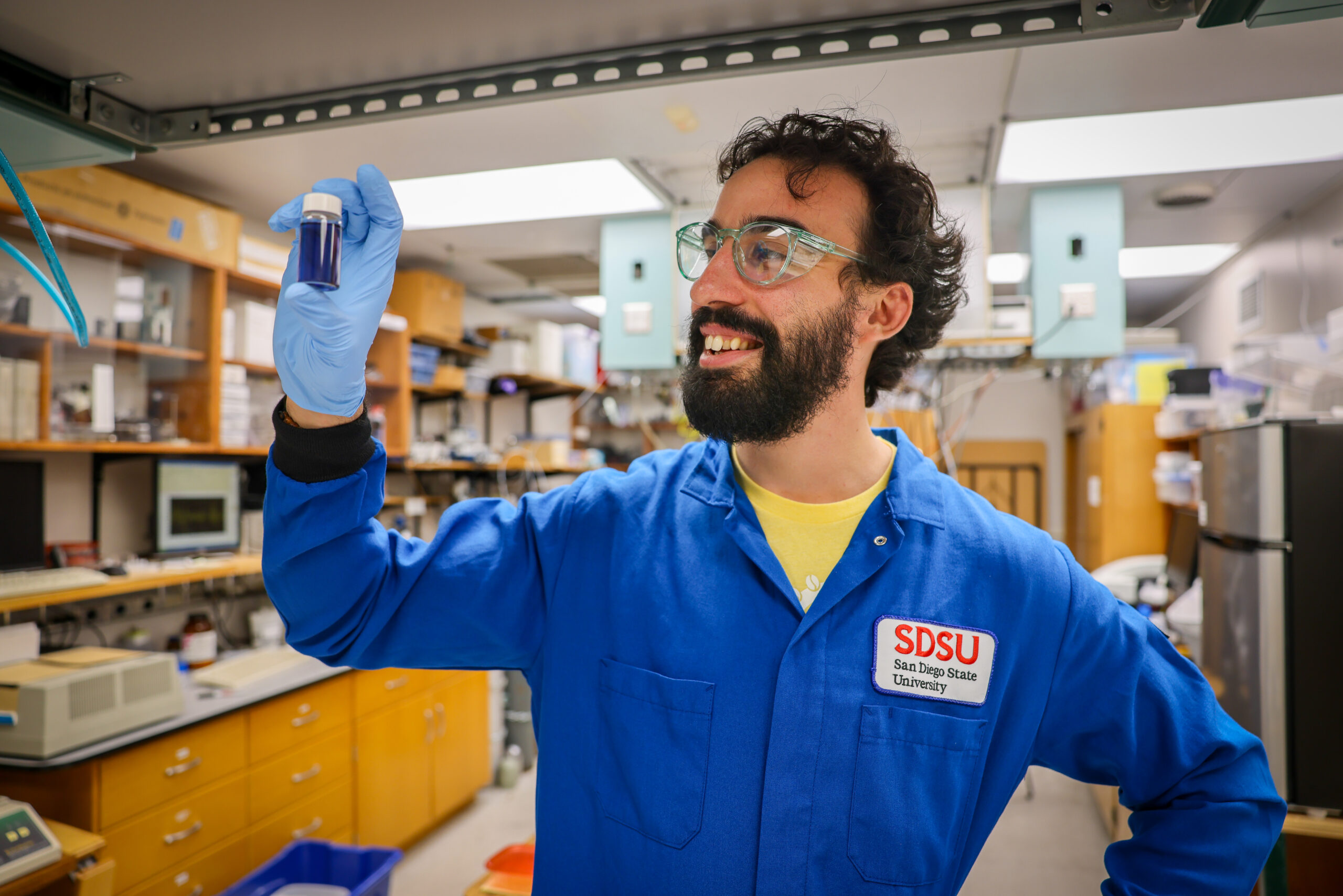 Male student wearing blue lab coat with SDSU logo holding up a small vile with blue liquid.